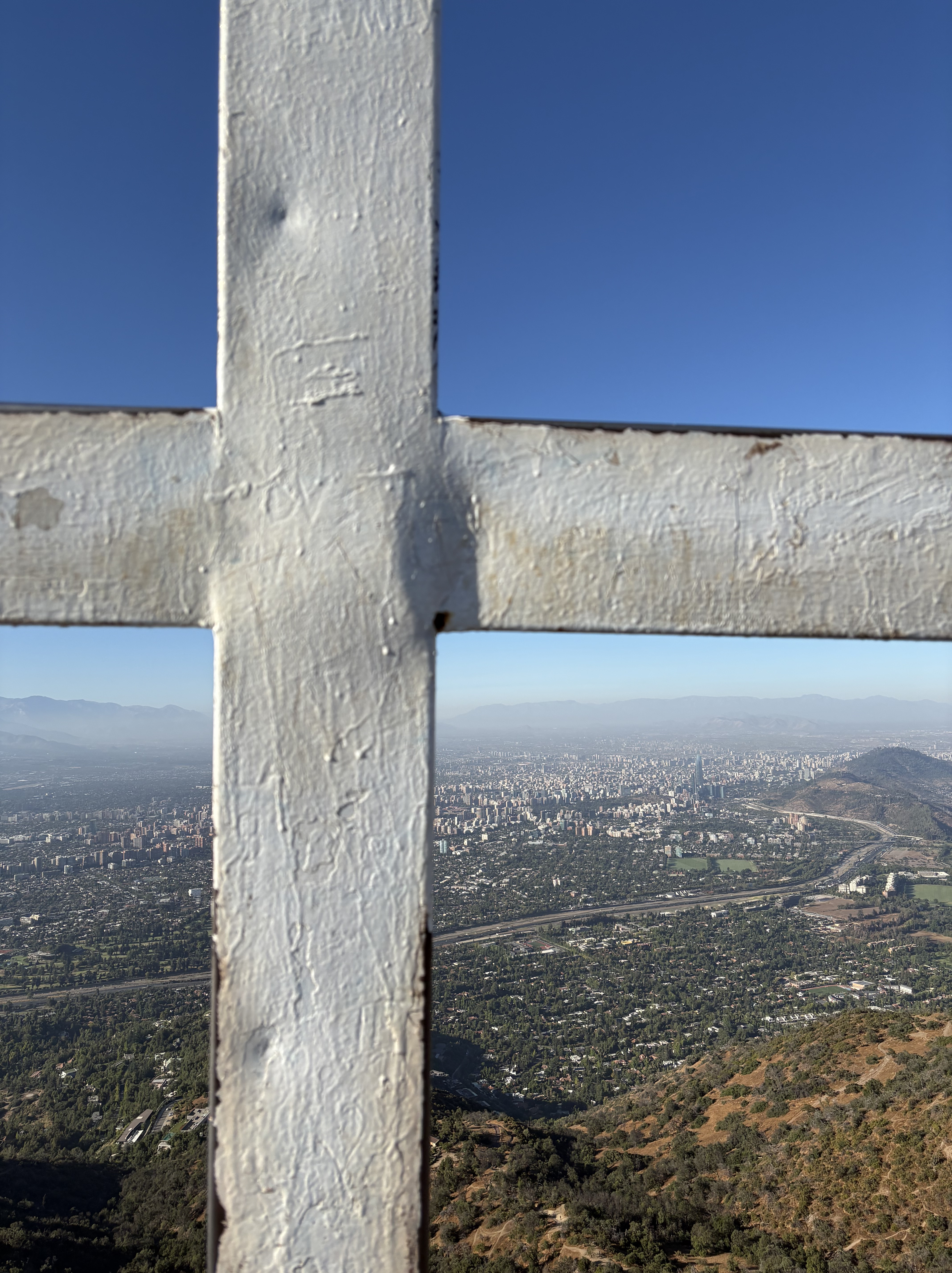 View of a city landscape framed by a white cross structure, overlooking greenery and urban buildings under a clear blue sky.